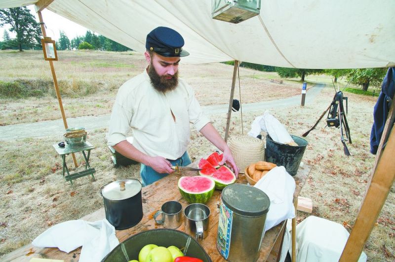 Marcus Larson/News-Register##Re-enactor Andrew Engel, portraying a soldier stationed at the Fort Yamhill garrison, works in the “kitchen,” slicing watermelon for the rest of his unit. The demonstration was part of a twice-yearly history day event at the state heritage area near Grand Ronde.