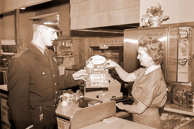 (January 7, 1958)##Making initial donation to a March of Dimes coin container, Mrs. Hazel Swinney places a coin in a can held by Police Chief Roy Brixey at Swinney’s Bakery. The container was one of nearly 100 containers and money cards distributed by the police department to McMinnville business firms for donation to the polio fund.