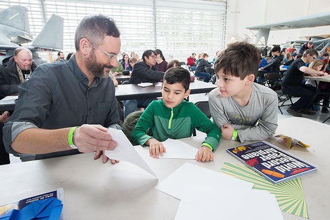 Marcus Larson / News-Register##Josh Ramseyer
shows his son Ansel, center, and his friend, Liam Tobin, the next step
in folding their own advanced paper airplanes.