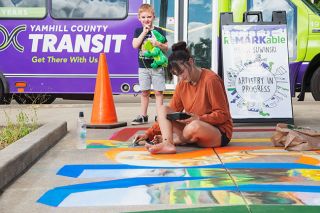Rusty Rae/News-Register##Caleb Johnson of McMinnville looks on Tuesday as artist M Lynn Suwinski works on the multi-image  TRANSIT  chalk mural at the Yamhill Transit plaza adjacent to First Street as part of Ridership Day.