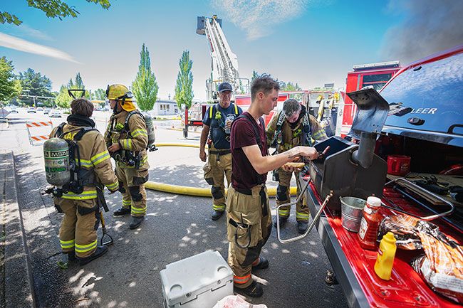 Rachel Thompson/News-Register##Isaiah Ellis from Dundee Fire takes a break for a hot dog. Behind Ellis are apparatus operator Doug Cummins, in ball cap, and Pete Goodman, self-described “captain with the volunteers,” both with McMinnville Fire.
