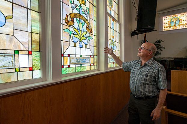 Rachel Thompson/News-Register ##Pastor Steve Hopper stands beneath stained-glass windows inside the Pioneer Evangelical Church sanctuary on Tuesday, reflecting on the history of both the windows and his tenure at the Dayton church. He is retiring after Sunday. Installed in the 1870s, the windows were funded by local donors, including a central pane honoring town founder Joel Palmer. “It’s the highlight window,” Hopper said. “It’s one piece, and it says ‘God is love’ — that kind of summarizes everything.” The flanking windows display the Greek symbols Alpha and Omega, symbolizing God as the beginning and the end.