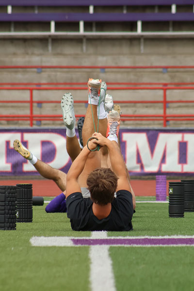 Nathan Ecker/News-Register##Linfield players lie on the ground and stretch their legs with bands at the start of practice on Aug. 14. Assistant coach Kernaghan emphasized proper movements and health during day one of fall workouts.