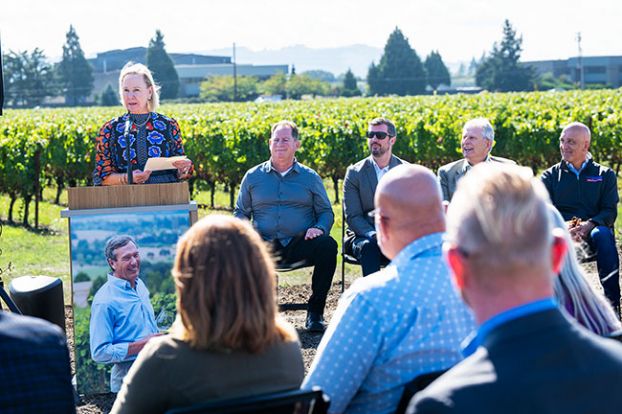 Rusty Rae/News-Register##LaRue Stoller talks about the vision her late husband, Bill Stoller, in photo on podium, had for the Evergreen campus and about how he wanted to benefit the community’s economy. She is speaking at the groundbreaking for the Evergreen Hotel, which will rise beside the vineyard and between the air museum and the water park. Beside Stoller are, from left, Patrick Nofield of Escape Lodging, which will run the Marriott AC hotel; Kraig O’Connor, president of R&H Construction; Stoller Group Chairman Wayne Marshall; and Sen. Bruce Starr, who also spoke at the groundbreaking.