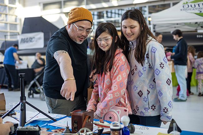 Rachel Thompson/News-Register##Jerome Castro of Beaverton gives his daughters, Elise, 11, and Aria, 14, a quick primer on film cameras, darkroom printing and the pre-smartphone tradition of photo albums at the Newberg Finds booth.