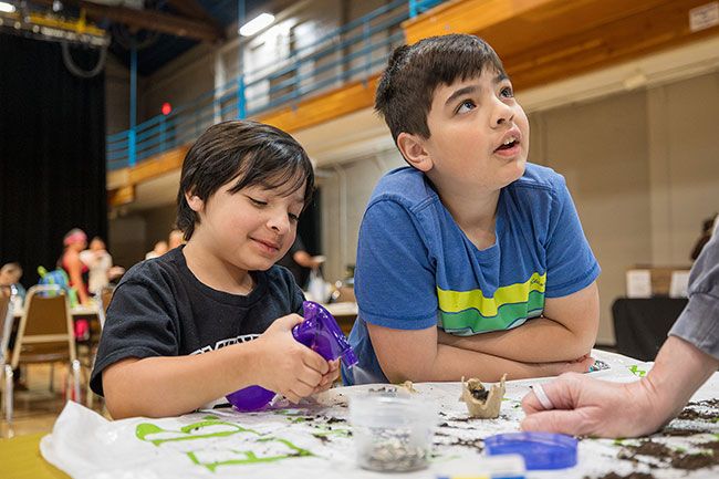 Rachel Thompson/News-Register##Aiden Pepinos, 7, and his brother Xavier, 11, plant sunflower seeds at the Edible Landscapes booth, where Xavier chats with Charlene Doland, president of Edible Landscapes.