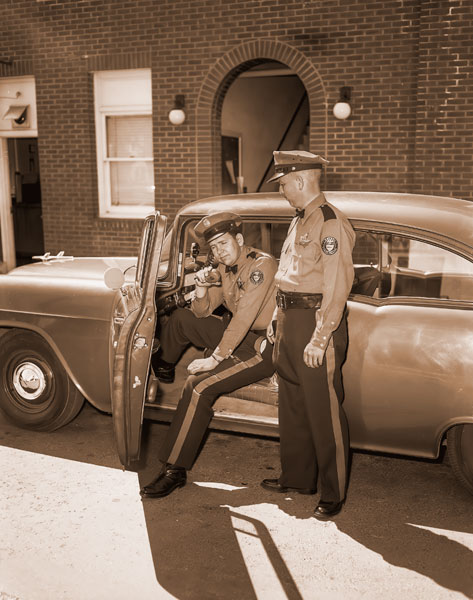 ## (May 2, 1956) The heat&rsquo;s on, but it won&rsquo;t make any difference to state police officers-they have changed to their light-weight summer uniforms. Gabardine shirts and slacks Tuesday replaced the heavy blouses, Sam Browne belt, breeches and boots. This is the first year the officers have worn slacks as part of their uniforms. Shown ready for work in their new uniforms are David Tindle, seated, and Glenn Shipman, from the McMinnville detachment of State Police.
