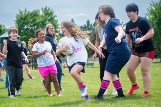 Emily Bonsant/News-Register##McMinnville girls rugby player Ofelia Patterson (center) carries the ball against her teammates and parents on June 6, at Joe Dancer Park as part of the closing of the Mid-Willamette Valley girls flag rugby season. (right) Sam Gamble and Vayda Ringen watch as Patterson rushes past the defense.
