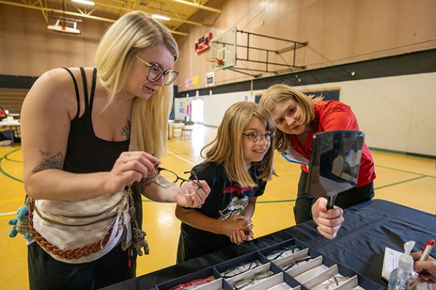 Rachel Thompson/News-Register##Emery Rivas, 8, looks at her reflection while choosing eyeglass frames with help from her mother, Kayla Lambert, left, and optometry student Katrina Billinton during a free eye clinic Friday, June 5, at Faulconer-Chapman School in Sheridan. After trying several options, Emery chose the pair in Lambert&rsquo;s hands. About 40 Sheridan students will receive prescription glasses through the clinic next month.