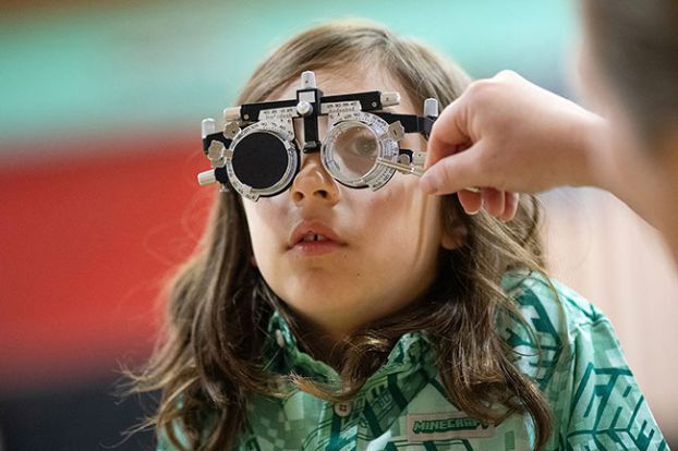 Rachel Thompson/News-Register##Emery Rivas, 8, looks at her reflection while choosing eyeglass frames with help from her mother, Kayla Lambert, left, and optometry student Katrina Billinton during a free eye clinic Friday, June 5, at Faulconer-Chapman School in Sheridan. After trying several options, Emery chose the pair in Lambert&rsquo;s hands. About 40 Sheridan students will receive prescription glasses through the clinic next month.