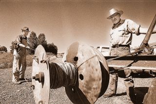 ##(July 21, 1976) Sam Bayliss (left) and Grant Youngberg operate a homemade winding device as they salvage telephone wire used by the Prune Center Telephone Company. Made from the rear axle of a car and driven from the tractor&rsquo;s PTO. Youngberg points out that it is also quite handy when working on fences. Founded in 1916, the company served an area east of Carlton. Before it shut down on May 1, it was one of the last farmer lines left in the county.